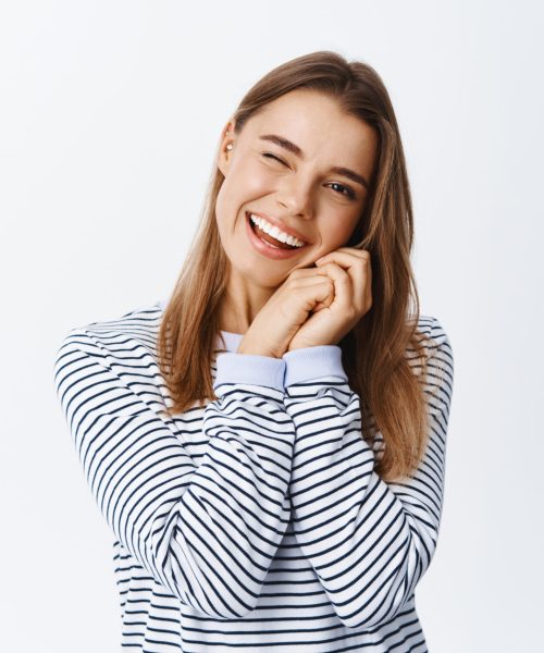 Cheeky young woman with blond hair, holding hands near natural face with light make up, winking and smiling at camera, standing over white background.